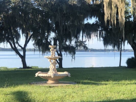 A decorative stone fountain is situated on a grassy area near a body of water, with large trees draped in Spanish moss in the background.
