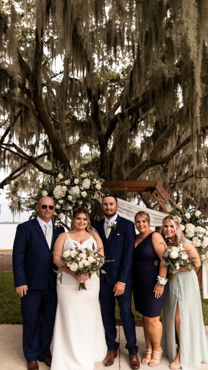 A group of five people dressed in formal attire stands together under a large, draping tree with Spanish moss, in front of a floral wedding arch at Starling Ranch Barn on the Lake in Fort Meade, FL.